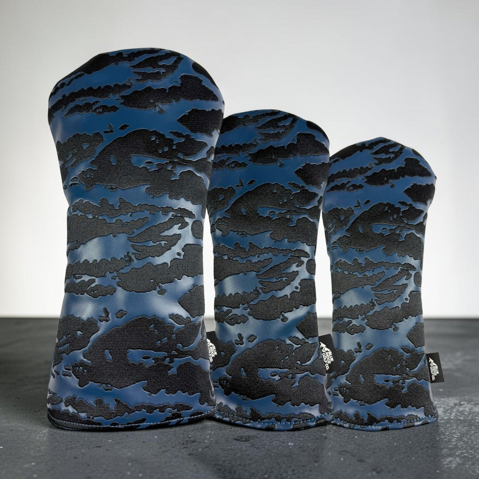 Three golf club headcover sleeves standing upright on a dark surface against a light gray background. The covers feature a blue and black abstract cloud-like camouflage pattern.