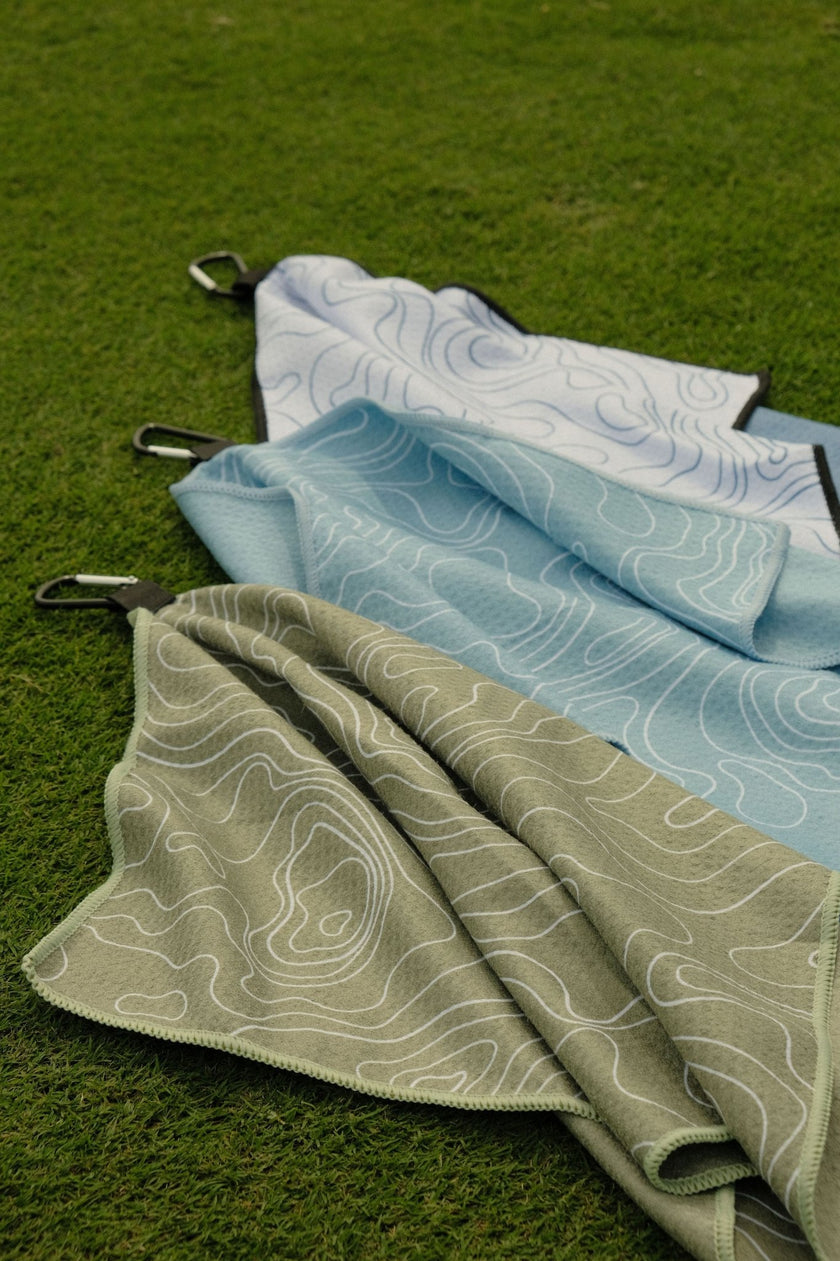 Folded golf towels in green, blue, and white on a grassy background