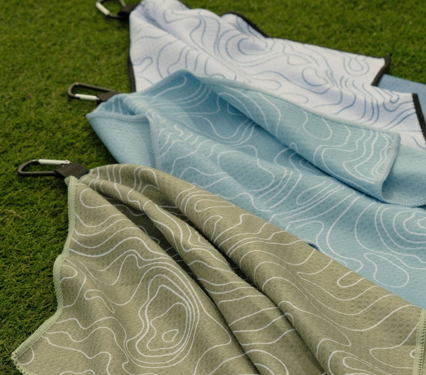 Folded golf towels in green, blue, and white on a grassy background