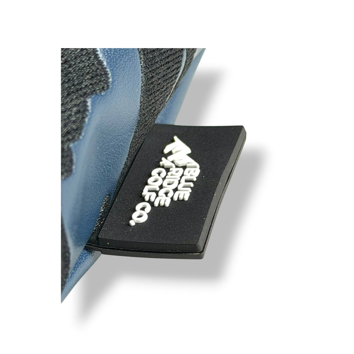 A close-up of a black rectangular patch with white stylized text and logo, attached to a blue and dark gray textured shoe or boot on a white background.